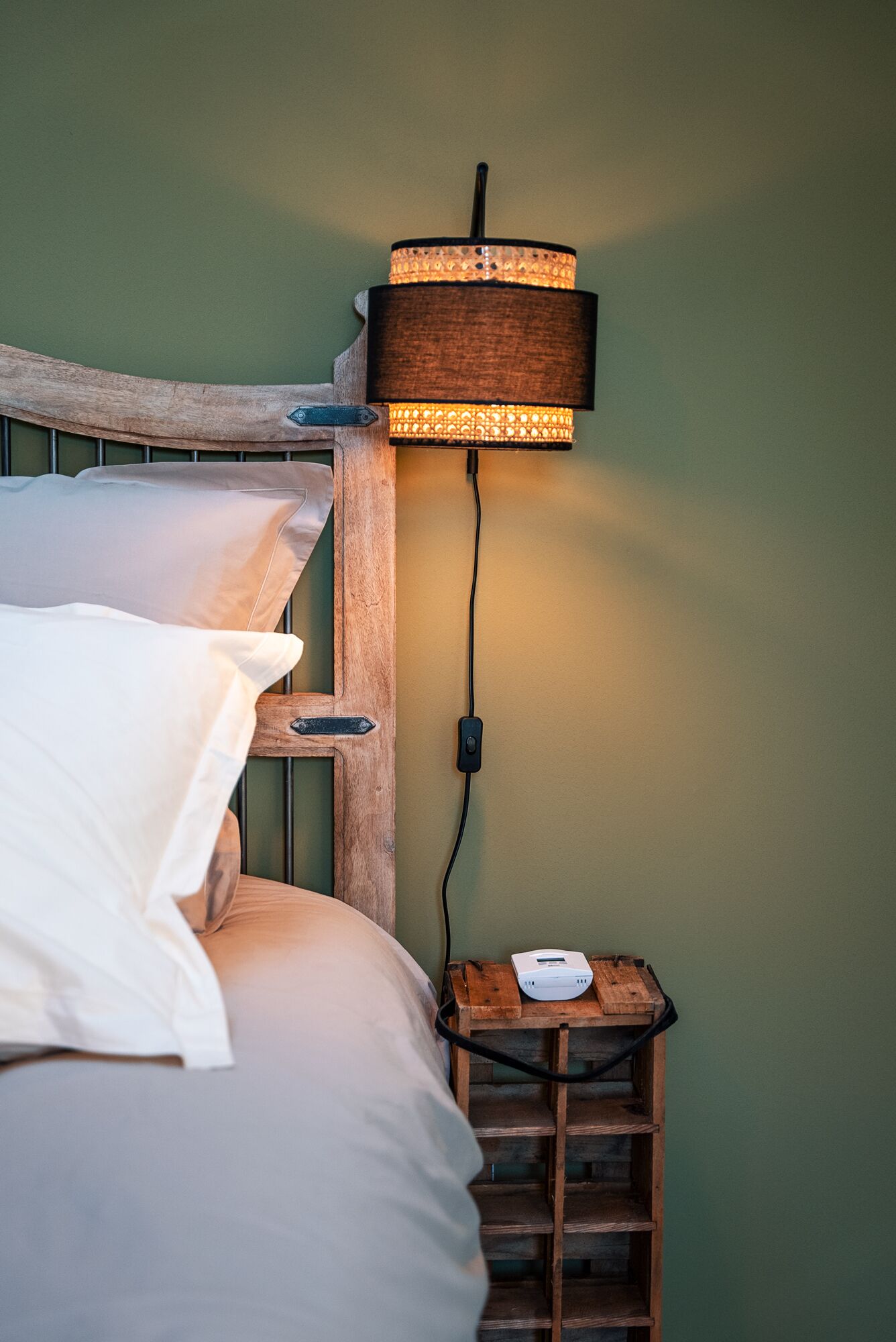 En säng med vita sängkläder, en sänggavel i trä, ett litet träbord vid sängen med en lampa och en grenuttag.