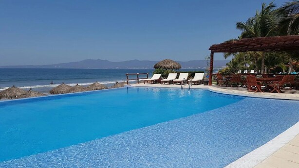 A clear blue pool with lounge chairs and a thatched structure, overlooking the ocean.