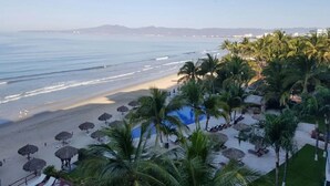 A beach resort with a pool, palm trees, and beach umbrellas.