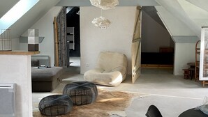 A spacious loft with high ceilings, exposed beams, and a skylight. There is a cozy seating area with a cushioned chair and ottoman, a dining table, and a cowhide rug. - Appartement Type Loft en Pleine Nature (Rumigny)