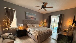 Une chambre avec un grand lit, un ventilateur de plafond, une table de chevet, une lampe, une fenêtre avec des rideaux, un tableau au mur et une vue sur l’extérieur.