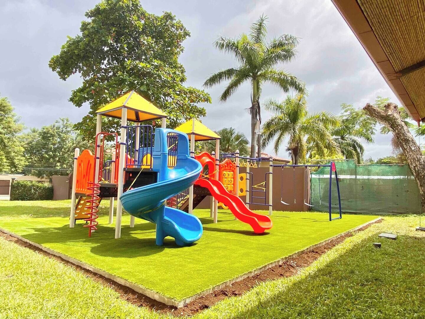 A playground with slides and climbing structures, surrounded by grass and palm trees.