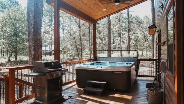 A wooden deck with a hot tub, grill, and a wooden railing overlooking a forest. - Stunning Farmhouse 1 mile to new casino sleeps 10 Hot Tub (Broken Bow)