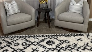 A living room with two grey armchairs, a small round table with a lamp and flowers, and a patterned rug.