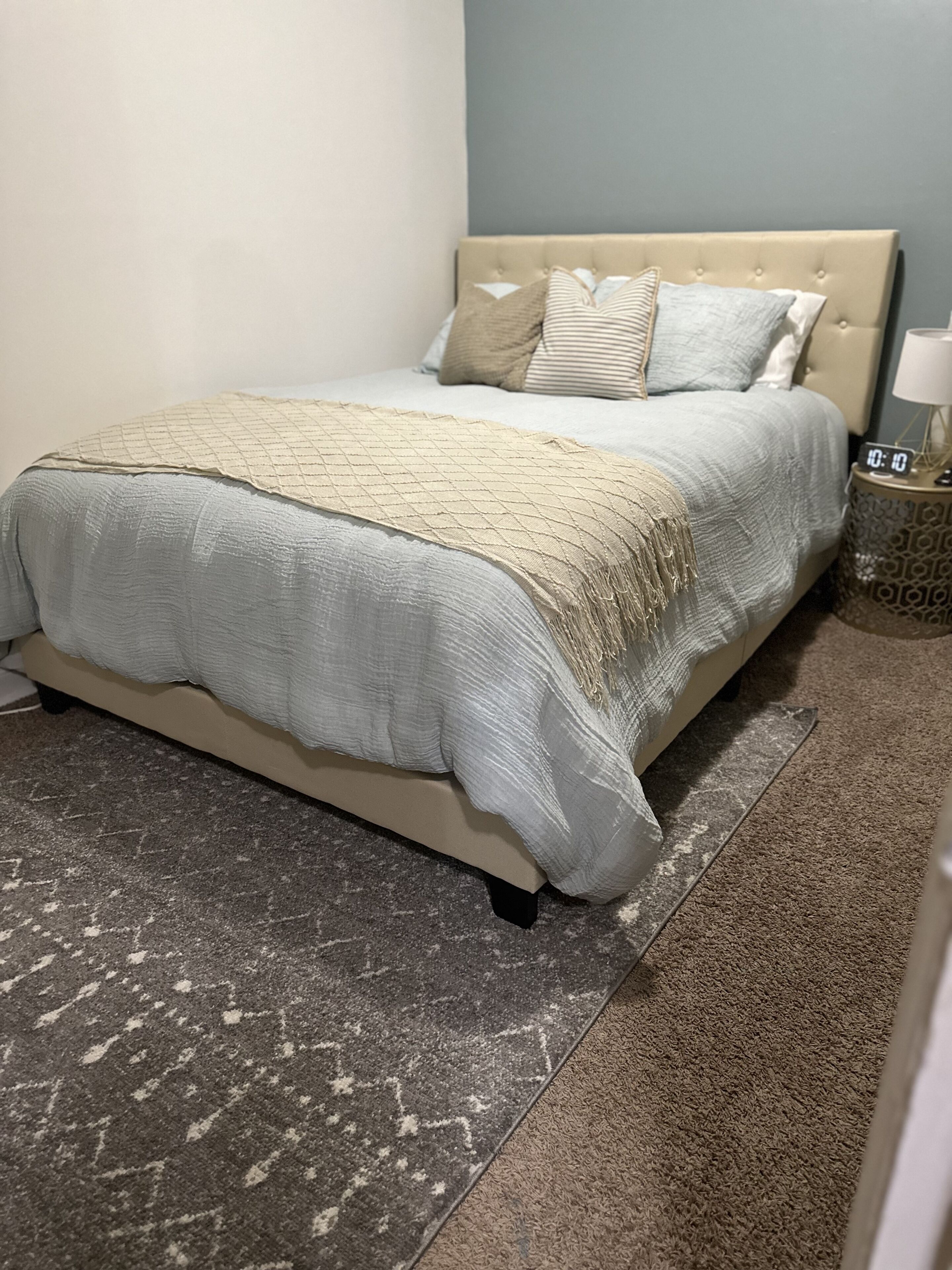 Een bed met een beige hoofdbord, een grijze spreidek en twee kussens. Er staat een klein nachtkastje met een lamp en een klok. De vloer heeft een tapijt met een patroon en een grijs vloerkleed.