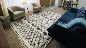 A living room with a beige sofa, a blue sofa, a patterned rug, a ceiling fan, and a dining area with chairs and a table.