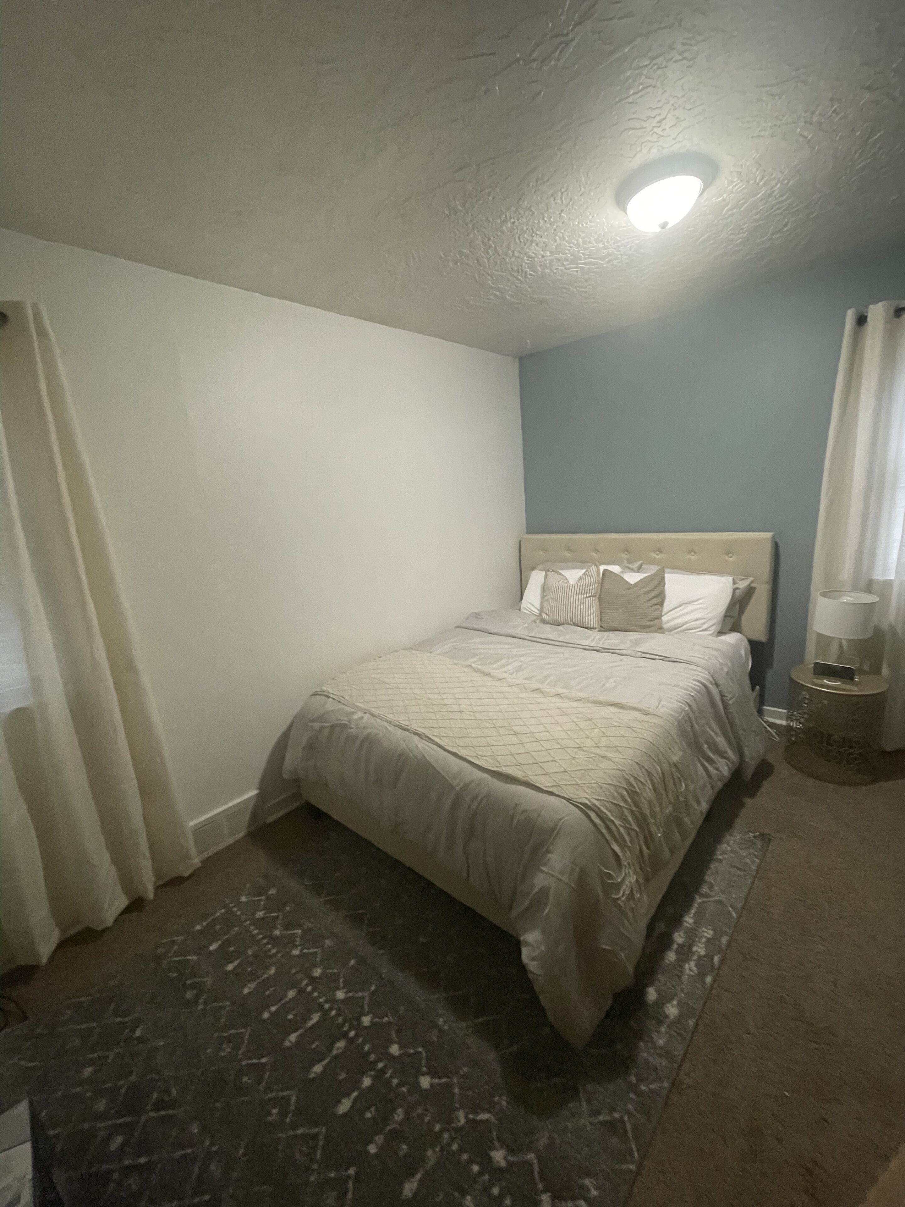 Een slaapkamer met een bed, nachtkastje, lamp, gordijn en plafondlamp.