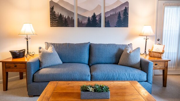 A living room with a blue sofa, two side tables with lamps, a wooden coffee table with a plant, and a wall with three mountain landscape paintings.