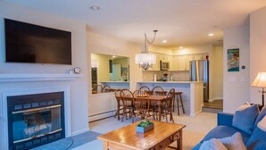 A living room with a fireplace, a TV, a dining area with chairs, and a kitchen in the background.