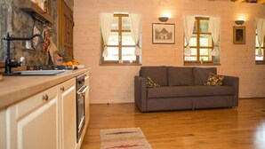 Ein gemütliches Zimmer mit Holzbalkendecke, einem grauen Sofa, einer Küchenzeile mit Herd und einem Fenster mit Vorhängen.