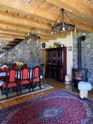 Cozy interior with a high ceiling, wooden beams, and a stone wall. The dining area features a wooden table and chairs, a patterned rug, and a dark cabinet with glass doors.