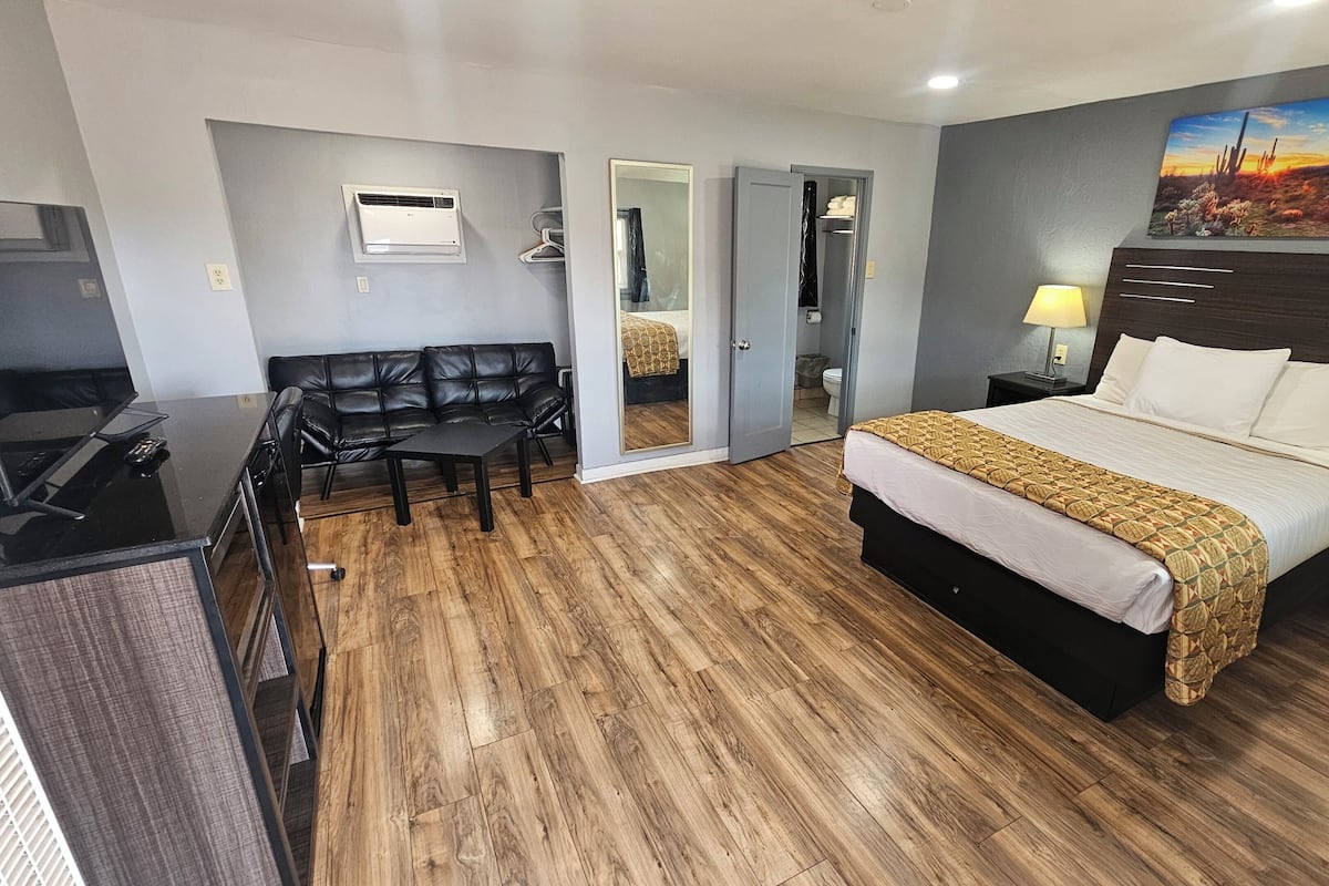 Une chambre d’hôtel avec un plancher en bois, un lit recouvert d’une literie blanche à motifs, un canapé en cuir noir, un bureau avec un ordinateur et un tableau accroché au mur.