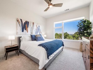 A bedroom with a bed, a nightstand, a dresser, a ceiling fan, a window with a view, and a painting on the wall. - Fully furnished luxury top floor condo across from Regis University (Denver)