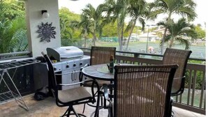 Eine Terrassenanlage im Freien mit Grill, Stühlen und Blick auf Palmen und einen Tennisplatz.