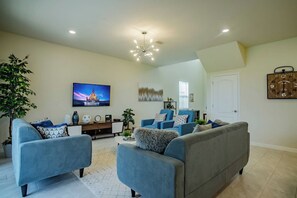 Un salon avec un canapé gris, des fauteuils bleus, un téléviseur à écran plat et un luminaire fixé au plafond.