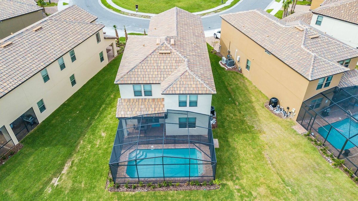 A residential area with a house featuring a swimming pool, surrounded by well-maintained lawns and neighboring houses.