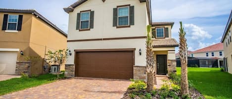 A two-story residential building with a brown garage door, a landscaped front yard with a palm tree, and a brick driveway.
