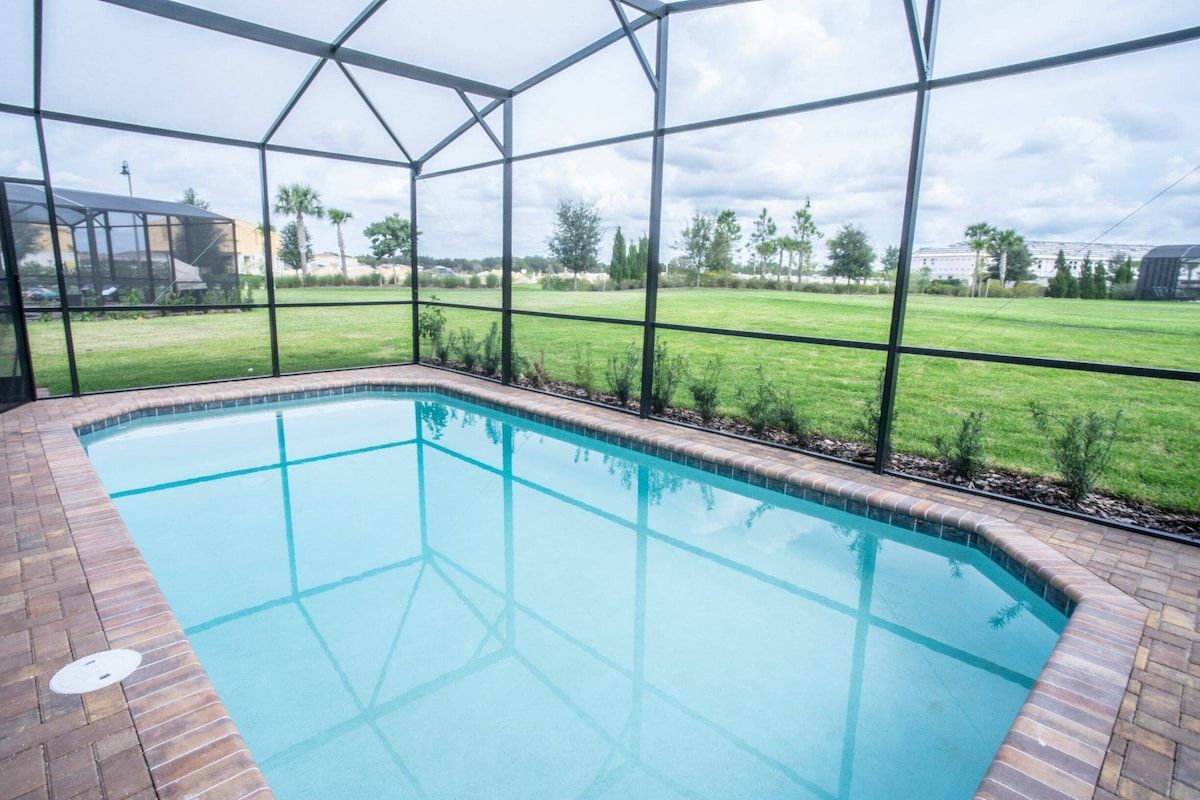 A swimming pool with a brick border, surrounded by a glass enclosure, and a view of a grassy field and trees.