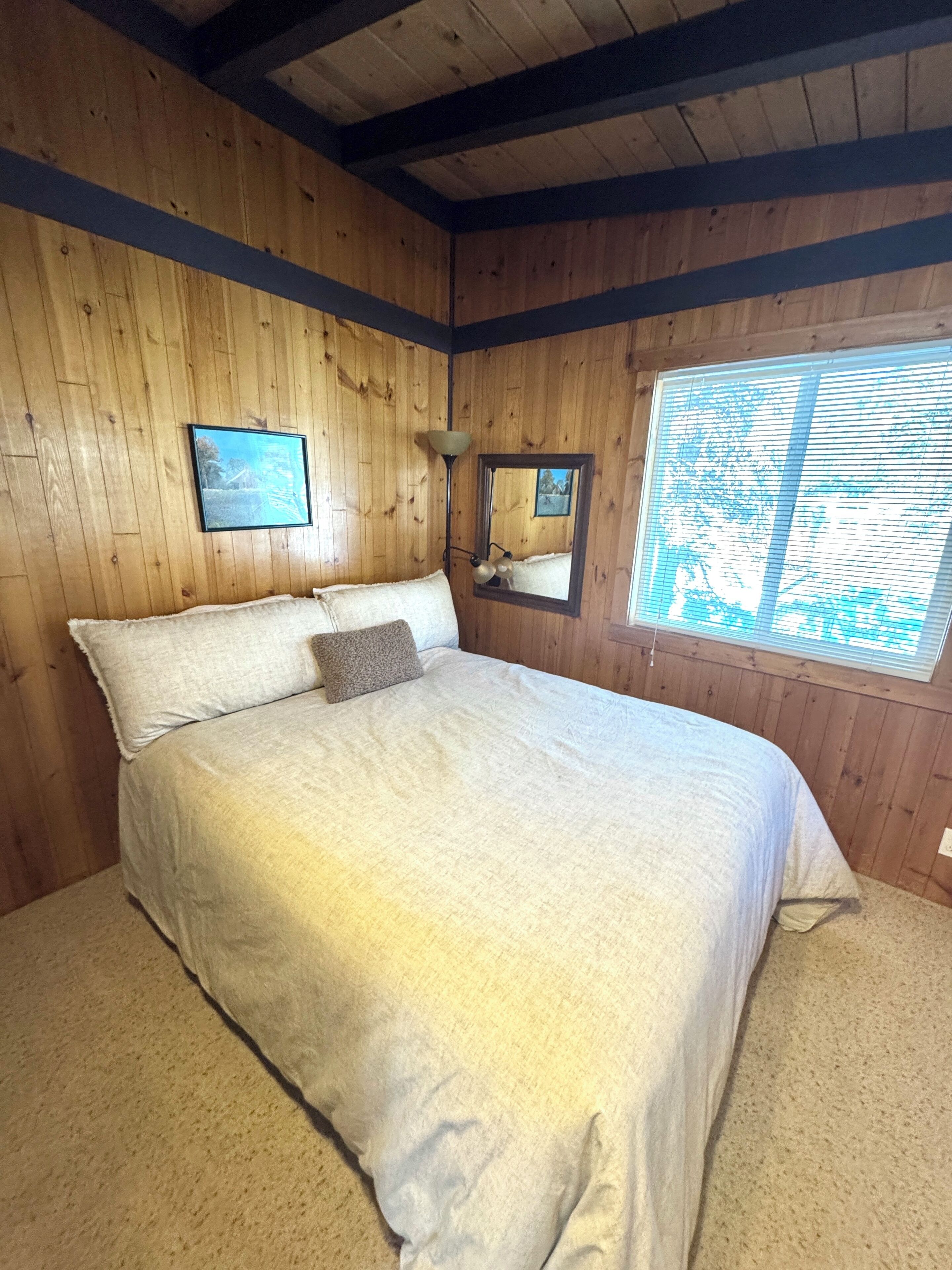 Ein Zimmer mit Holzwandverkleidung, einem Bett, einem gerahmten Bild an der Wand, einem Spiegel und einem Fenster mit Jalousien.