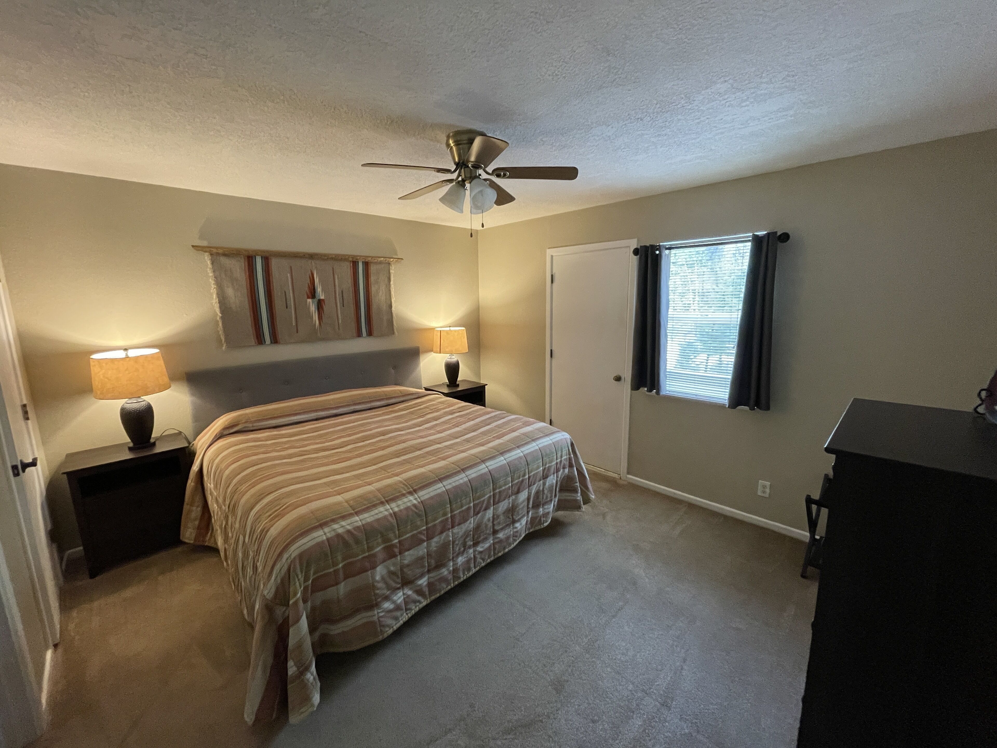 A bedroom with a bed, two bedside tables with lamps, a ceiling fan, a door, and a window with curtains.