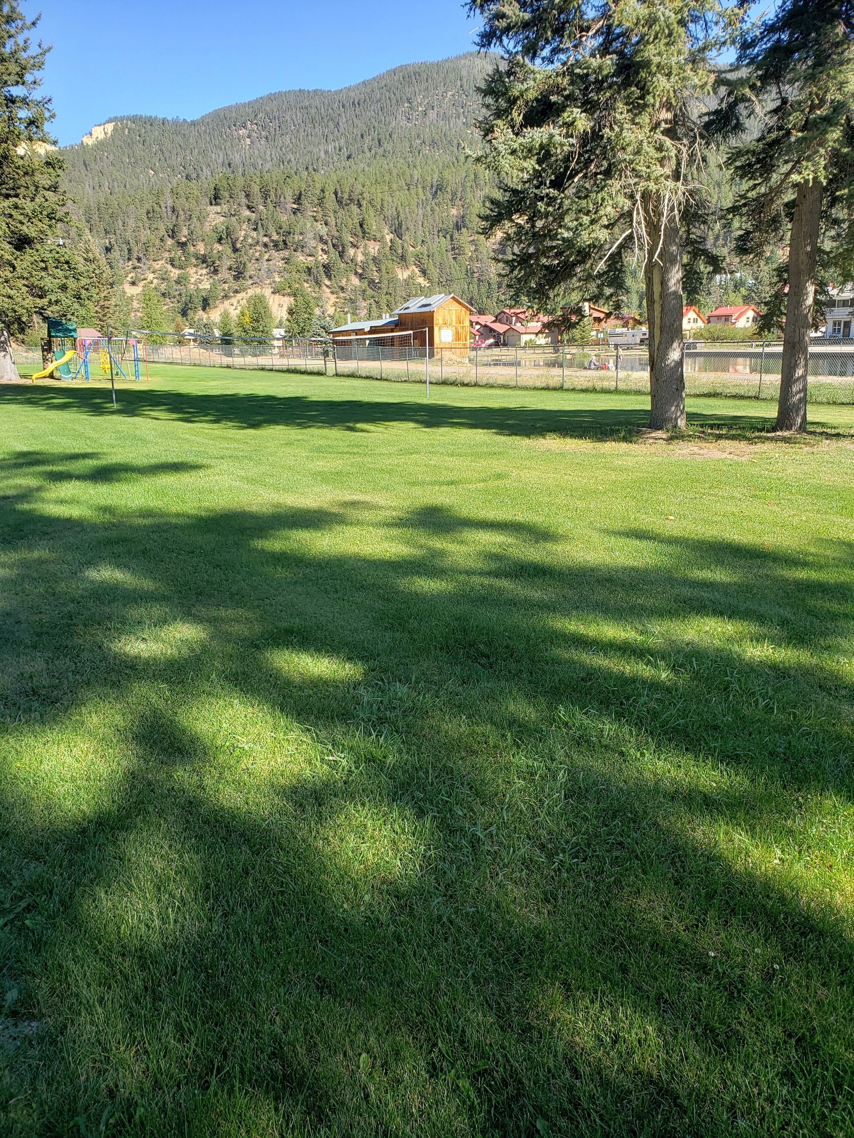Un área de parque con césped, un parque infantil, una estructura de madera y una montaña al fondo.