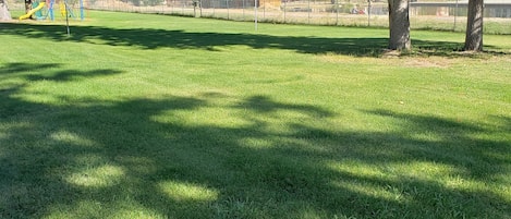 Un área de parque con césped, un parque infantil, una estructura de madera y una montaña al fondo.