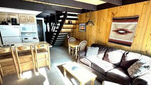 A cozy living room with a wooden ceiling, a sofa, a coffee table, and a kitchen area with a refrigerator and stove. - Auslander Condominiums (Red River)