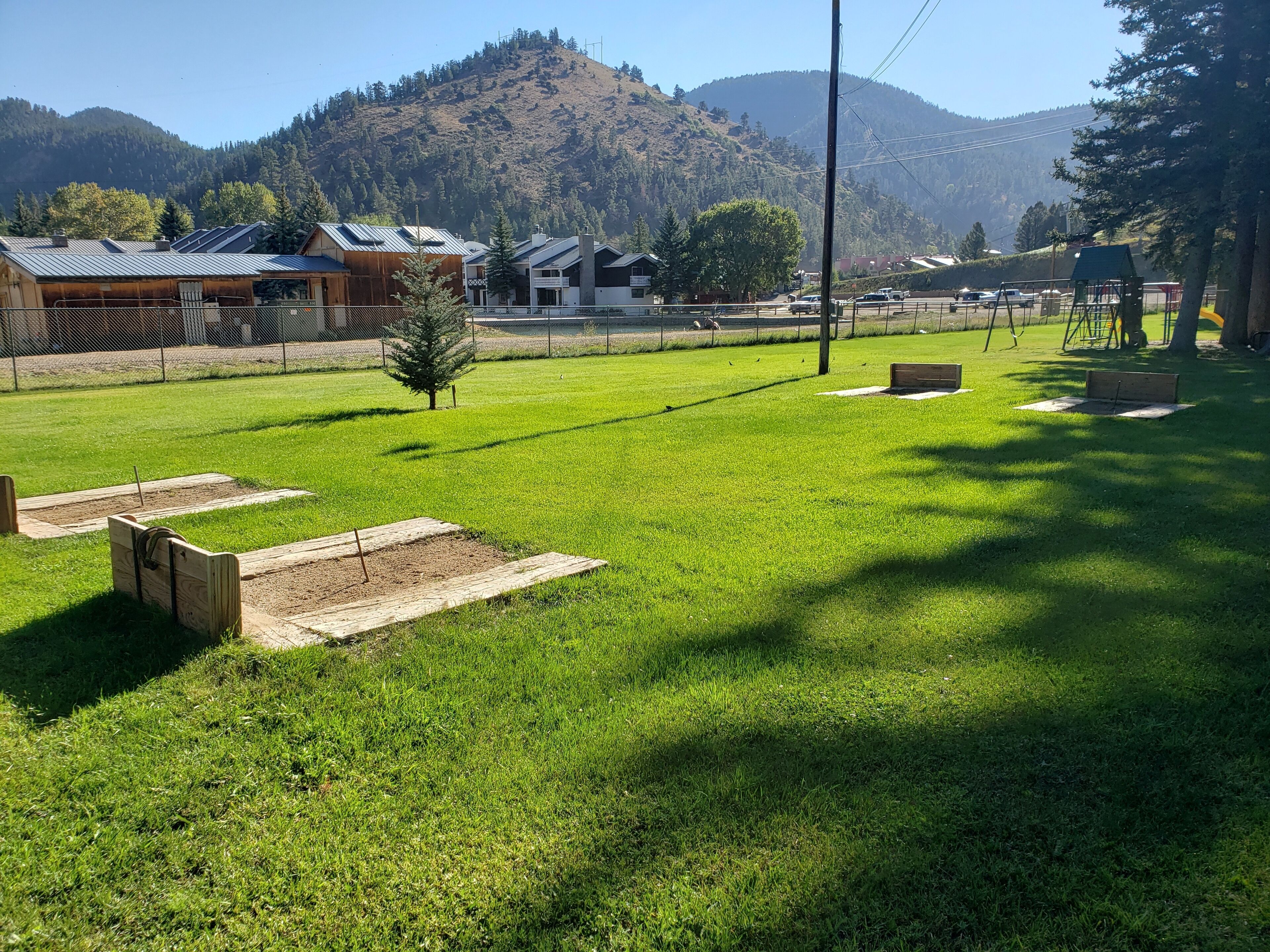 A grassy field with wooden benches and a small tree, surrounded by buildings with metal roofs and a mountain in the background.