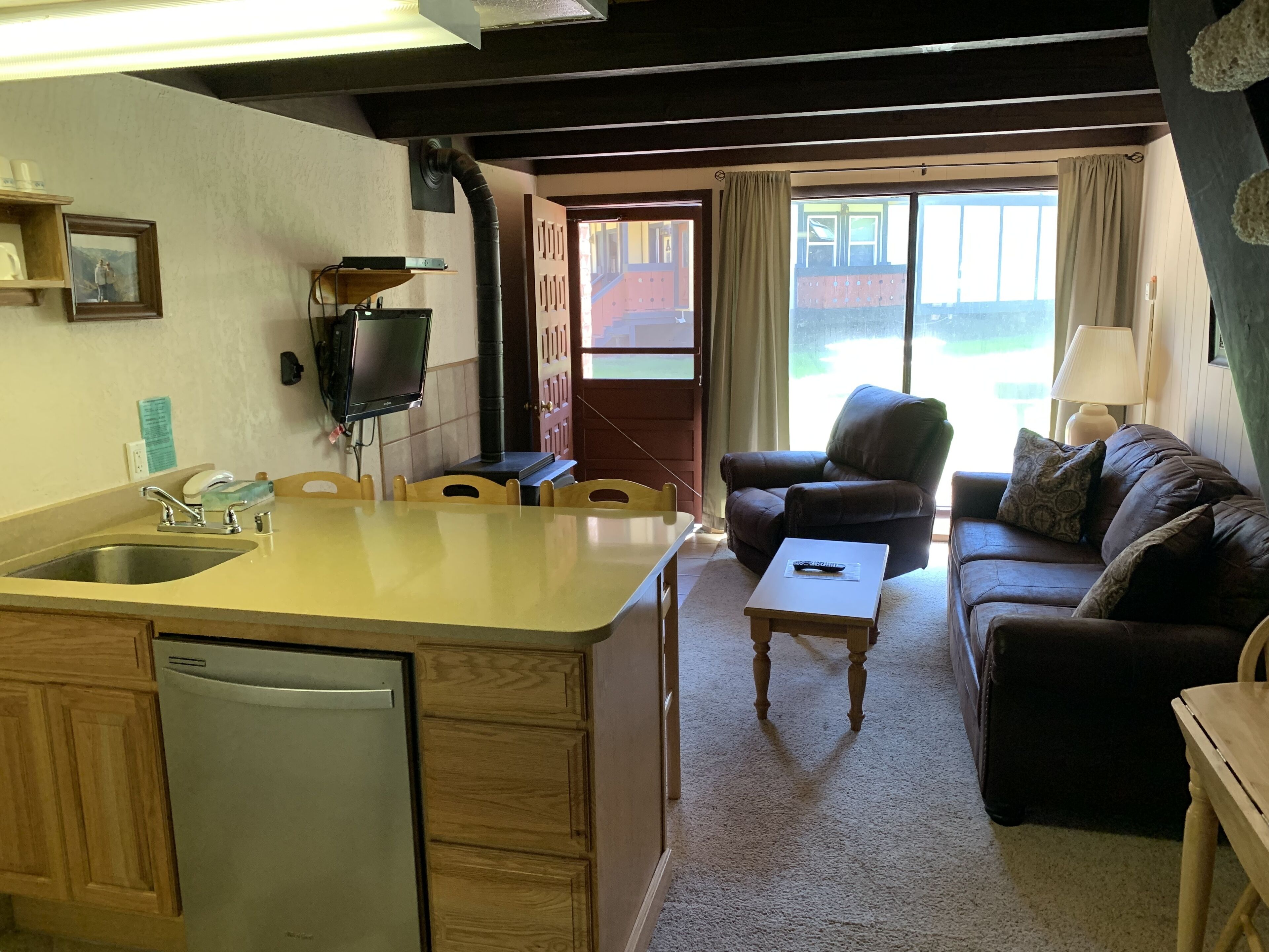 A compact kitchen with a sink, stove, and a television. There is a dining area with a table and chairs, and a living room with a sofa and a lamp.