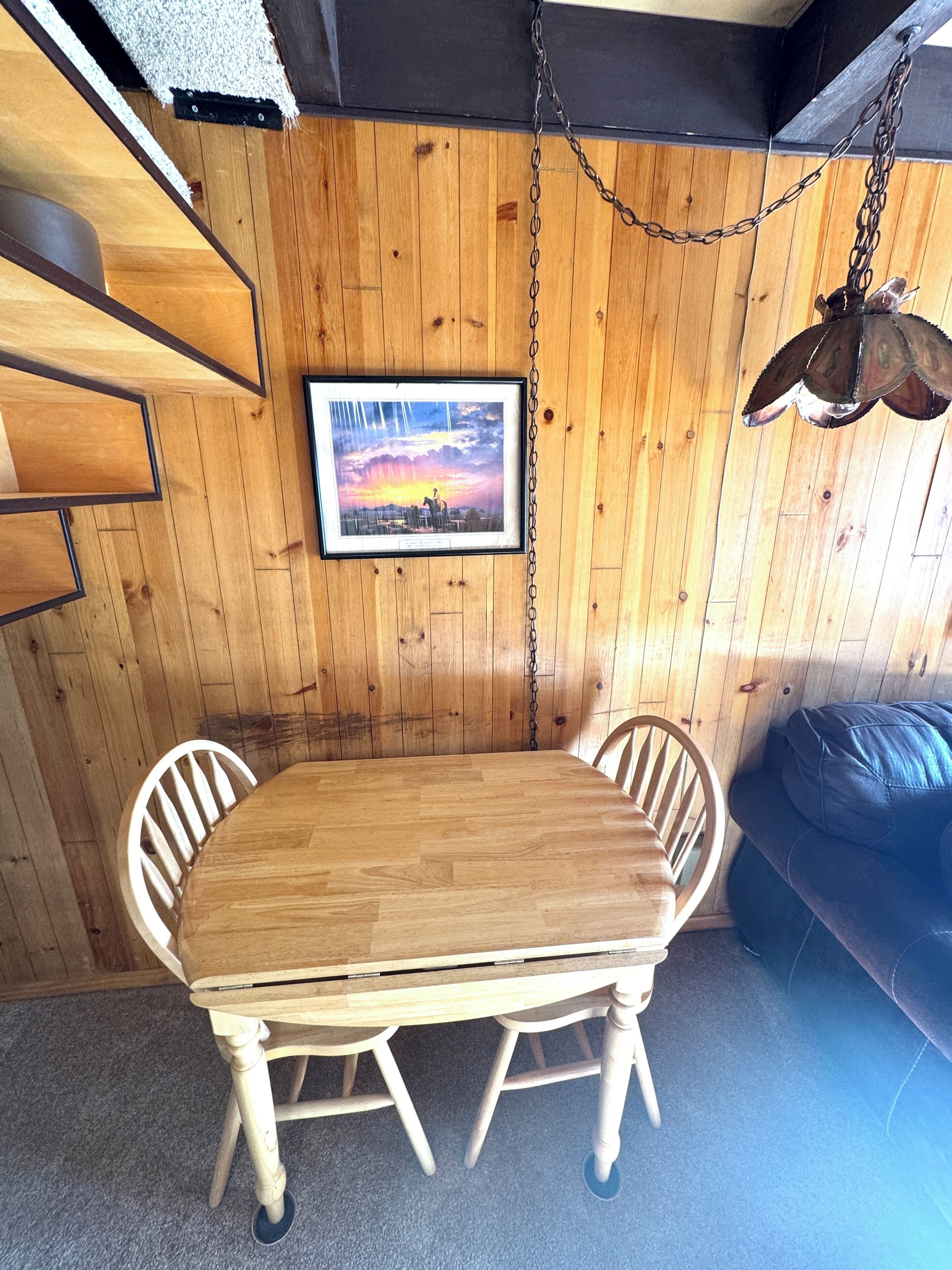 A wooden dining table with chairs, a hanging light fixture, and a framed picture on the wall.