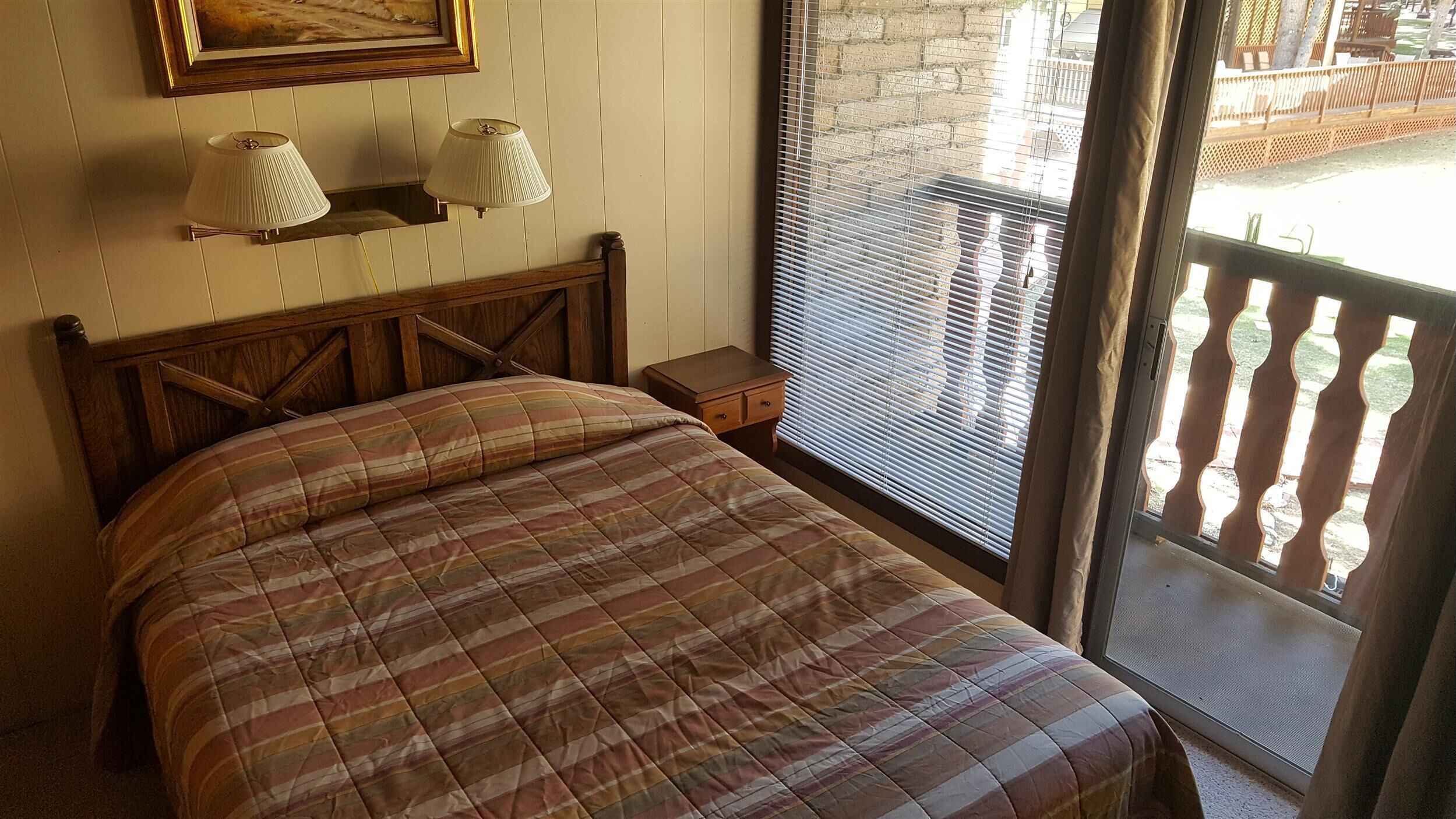 Une chambre à coucher avec un lit, deux lampes fixées au mur, une table de chevet en bois et une fenêtre avec des stores.