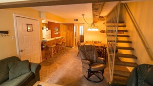 A living room with a sofa, a recliner, a dining area with chairs, a kitchen in the background, and a staircase. - Auslander Condominiums (Red River)