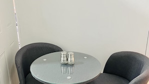 Et rundt bord med to grå polstrede stole, to salt- og pebershakere, samt en hvid væg med en lyskontakt.