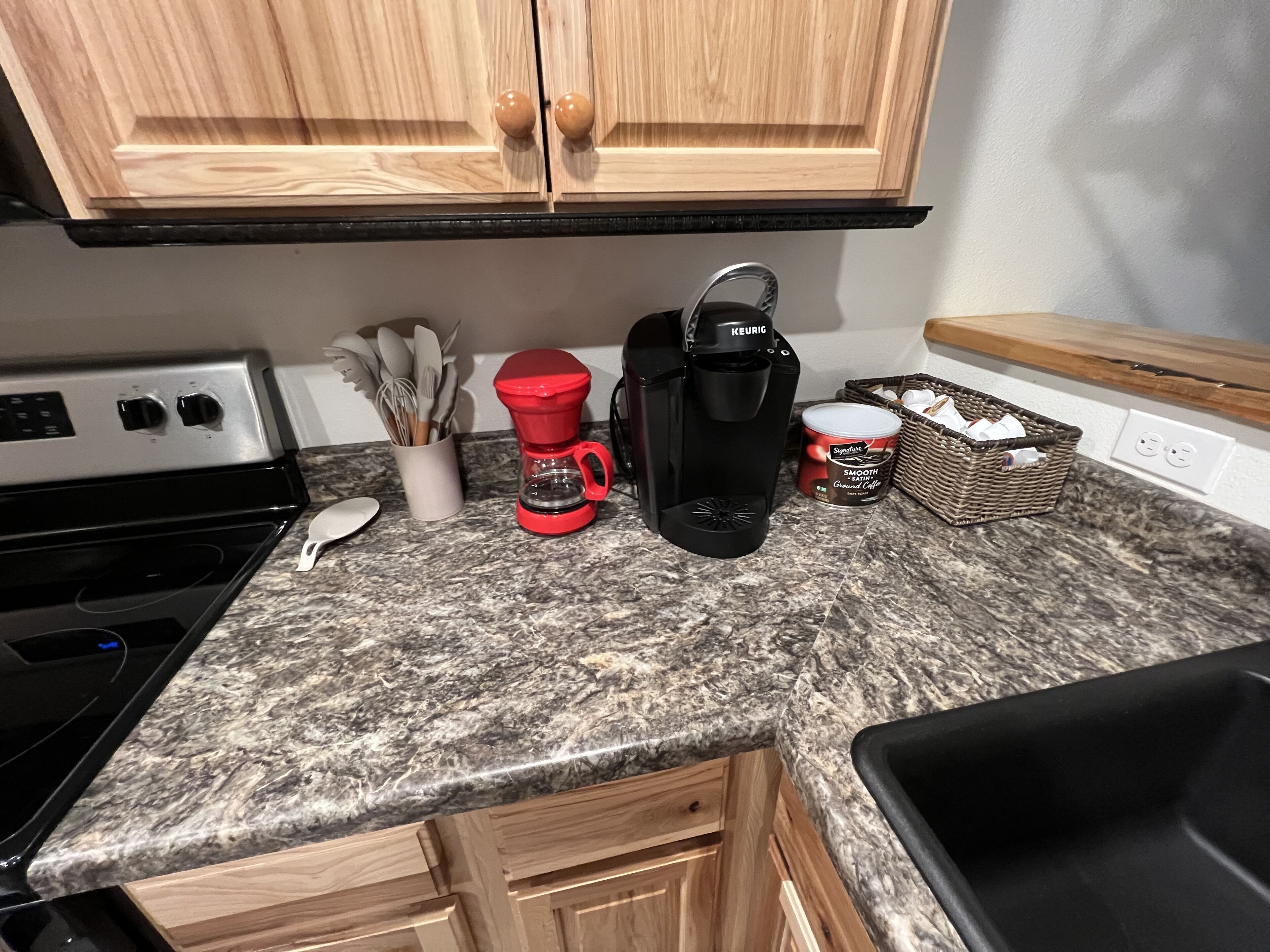 A kitchen with granite countertops, wooden cabinets, a coffee maker, a basket, and a can of coffee.