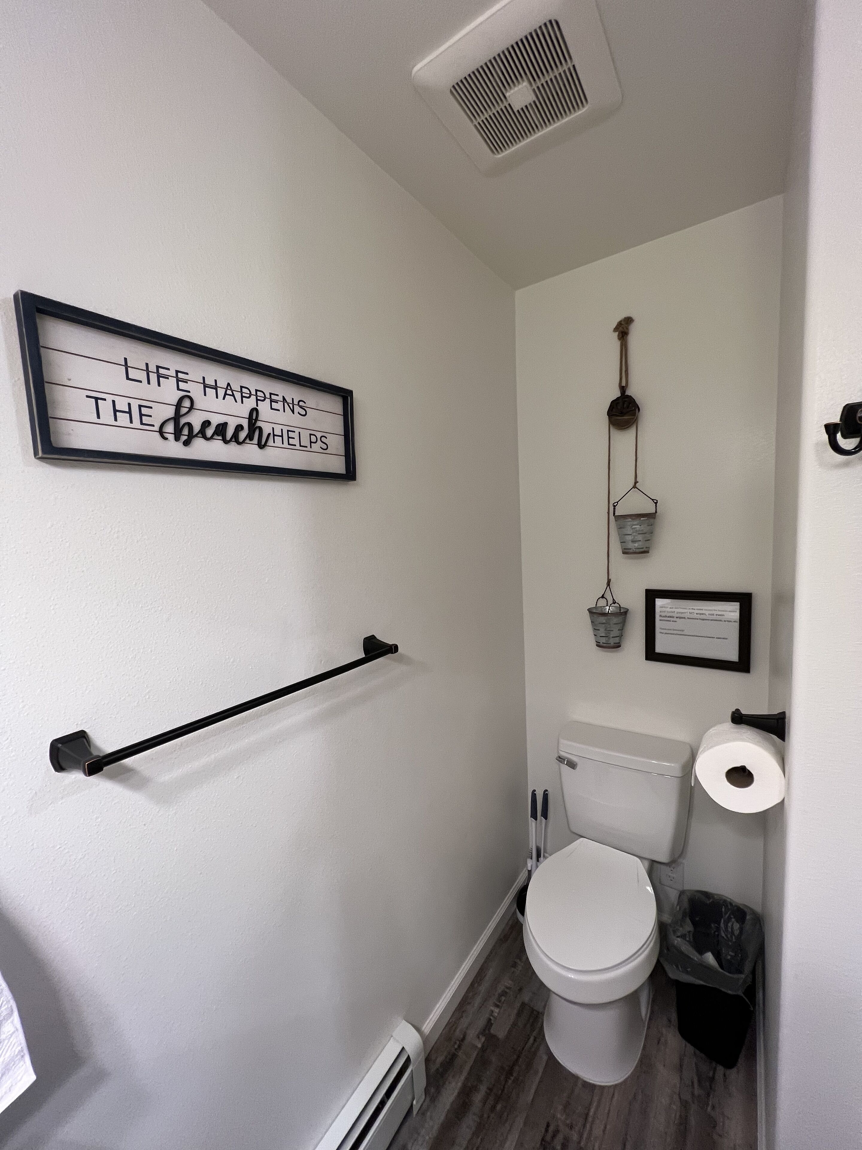 A small, narrow bathroom with a toilet, a towel rack, a wall-mounted sign, and a small bucket hanging on the wall.