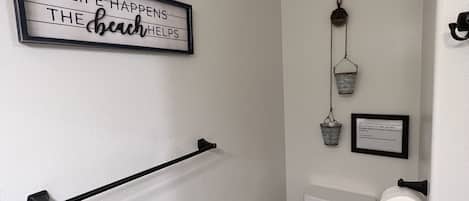 A small, narrow bathroom with a toilet, a towel rack, a wall-mounted sign, and a small bucket hanging on the wall.