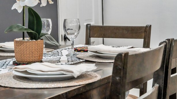 A dining table set with white plates, napkins, and a glass. There is a woven basket with a plant and a glass on the table.