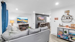 A modern living room with a grey sofa, a TV, a dining area, and a bookshelf with board games.