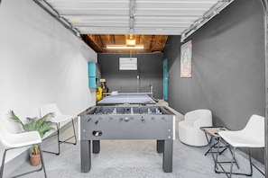 Une pièce avec un baby-foot, des chaises blanches, une petite table et une plante en pot.
