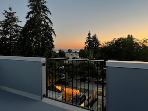 Un balcón con barandilla de metal que da a una zona residencial, árboles y una puesta de sol.