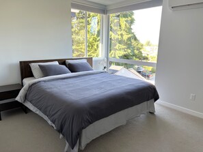 Un dormitorio con un ventanal grande, una cama con ropa de cama gris y una mesita de noche.