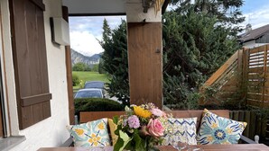 A wooden table with a vase of flowers and wine glasses, set against a white wall with a window overlooking a garden and mountains.