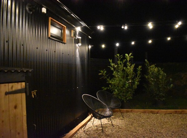 A dark metal building with a wooden door, a window, and a string of lights. There are two chairs outside.