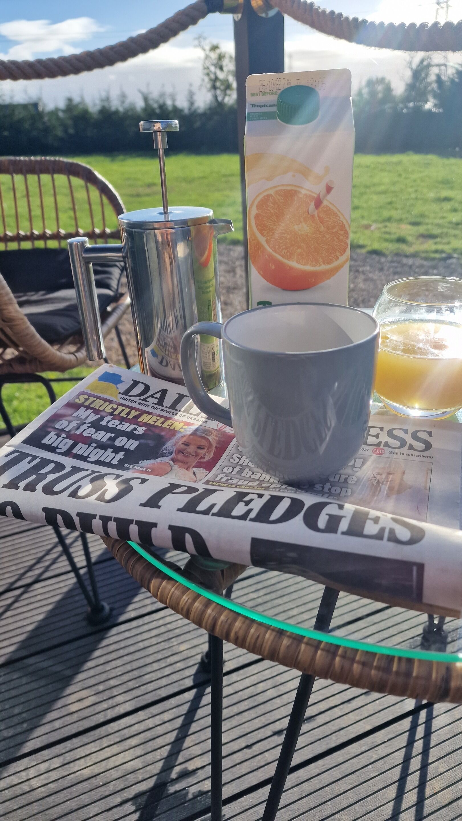 Una mesa con periódico, cafetera, taza, vaso de jugo y cartón de jugo de naranja en una terraza con vistas al exterior.