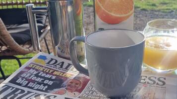 Uma mesa com jornal, cafeteira, caneca, copo de suco e um recipiente de sumo de laranja numa varanda com vista para o exterior.