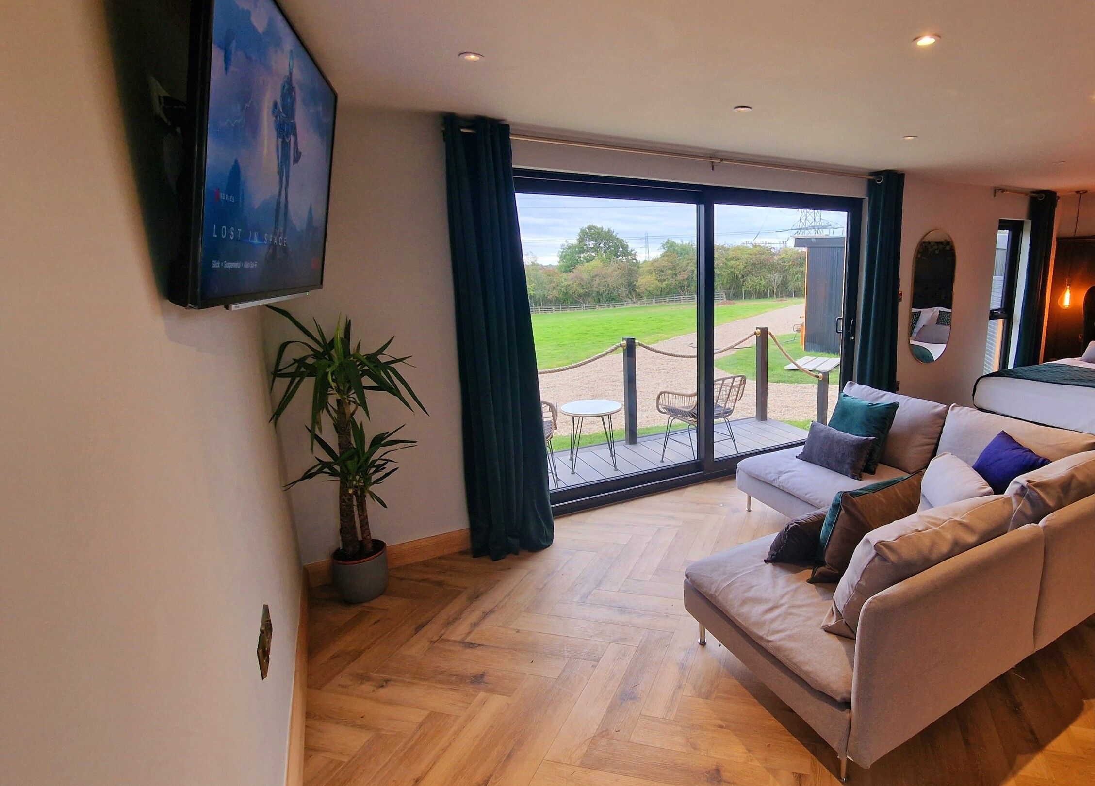 A modern bedroom with a large bed, a sofa, and a TV. There is a balcony with a view of the outdoors.