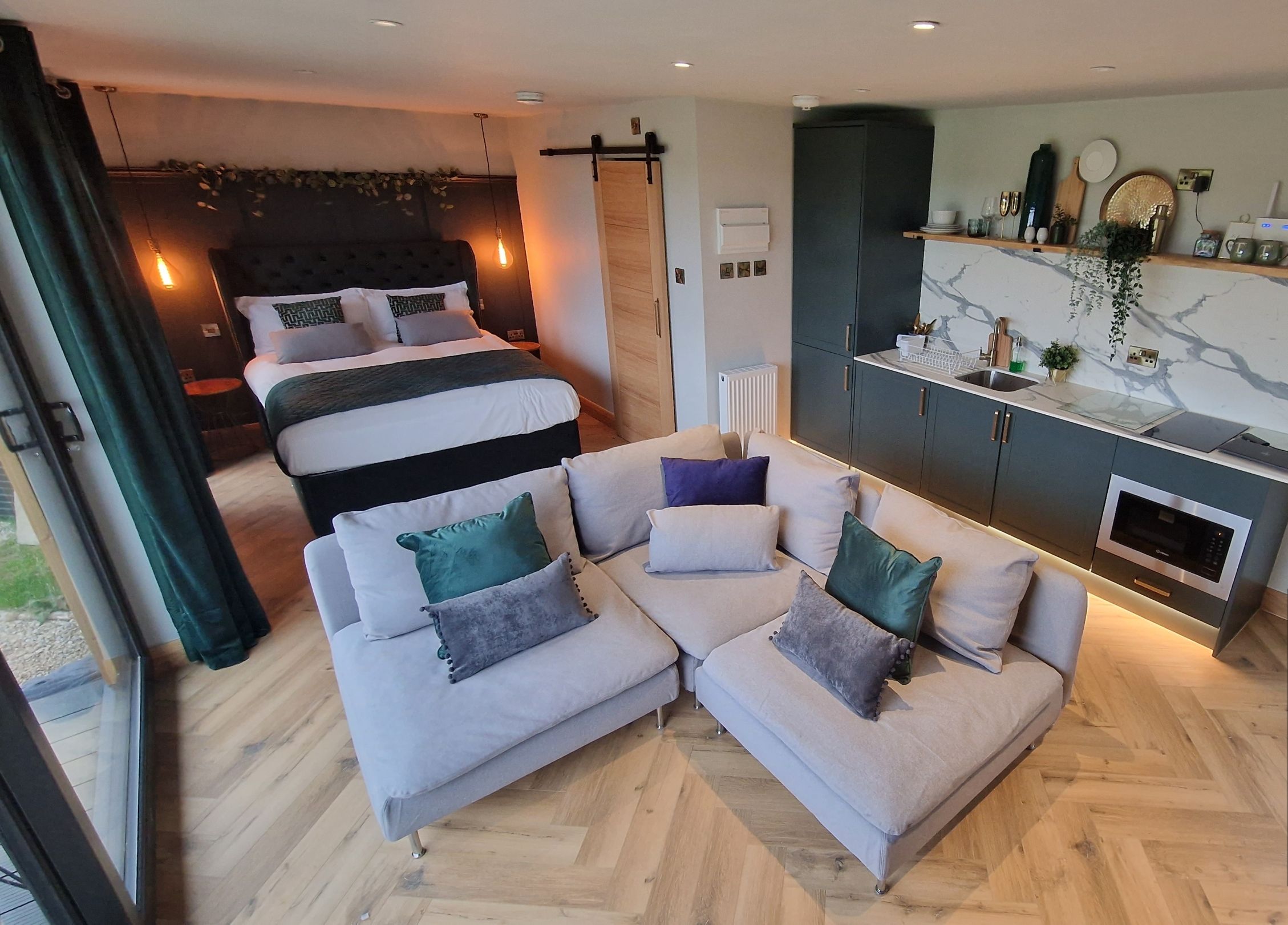 Ein modernes Hotelzimmer mit einer Sitzecke, einem Bett mit dunklem Kopfteil, einer Küchenzeile mit integriertem Backofen und einem großen Spiegel an der Wand.