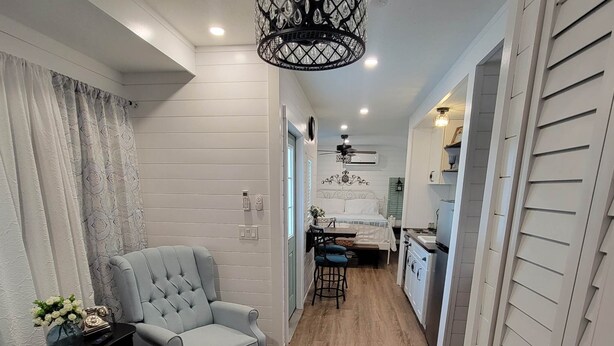A compact living space with a grey armchair, a small round table, and a kitchen area with white cabinets and a black ceiling light fixture. - Romantic Escape on Peaceful Mini Farm (Coleman)
