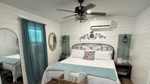 A bedroom with a bed, a ceiling fan, a mirror, a window with curtains, and a wall-mounted air conditioner. - Romantic Escape on Peaceful Mini Farm (Coleman)