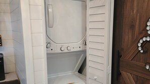 A compact laundry room with a front-loading washer and dryer, a small shelf, and a wooden door with decorative elements. - Romantic Escape on Peaceful Mini Farm (Coleman)